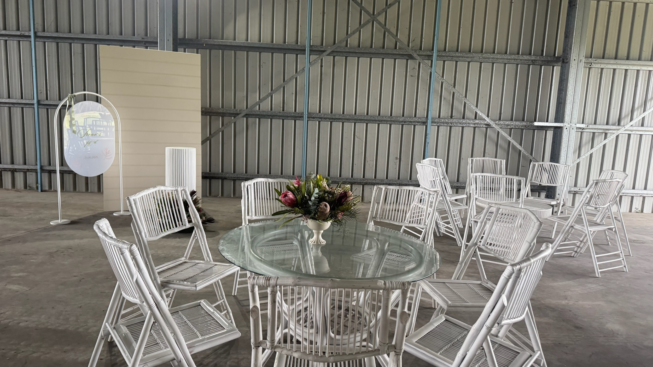 A round glass table with a floral centerpiece surrounded by white wicker chairs inside a large industrial warehouse or event space with metal walls.