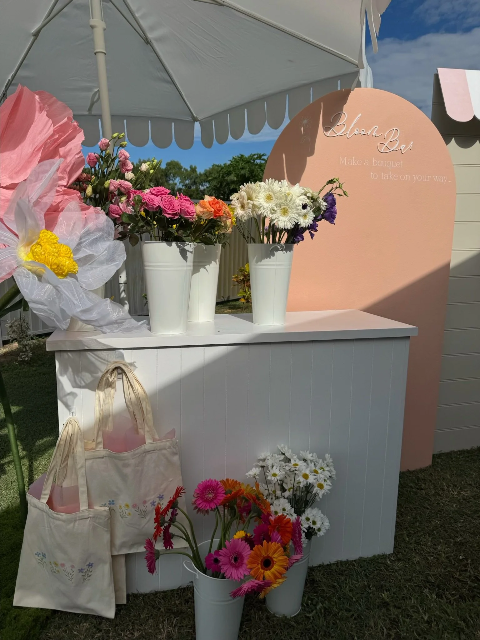 Display of colorful flowers in white vases and buckets with a pink sign that reads 'Blossom Bar: Make a bouquet to take on your way,' under a white parasol outdoors on a sunny day.