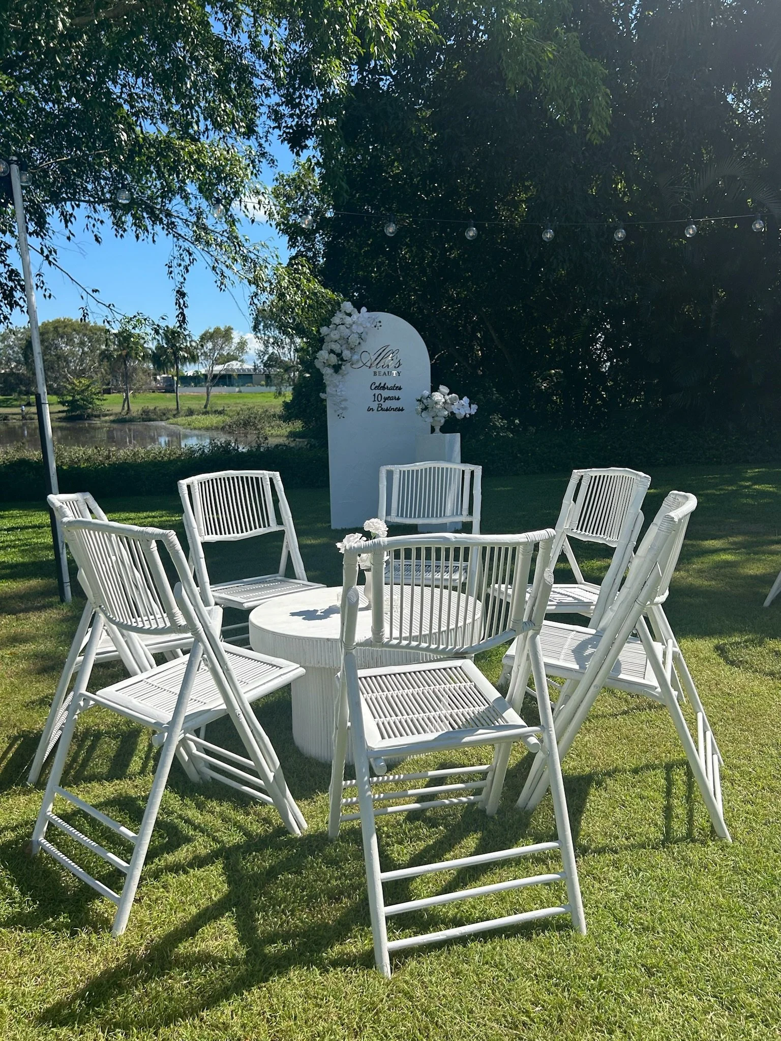 Outdoor event setup with a round white table surrounded by six white chairs on a grassy area. In the background, there is a white sign decorated with flowers, and trees over a pond or lake.