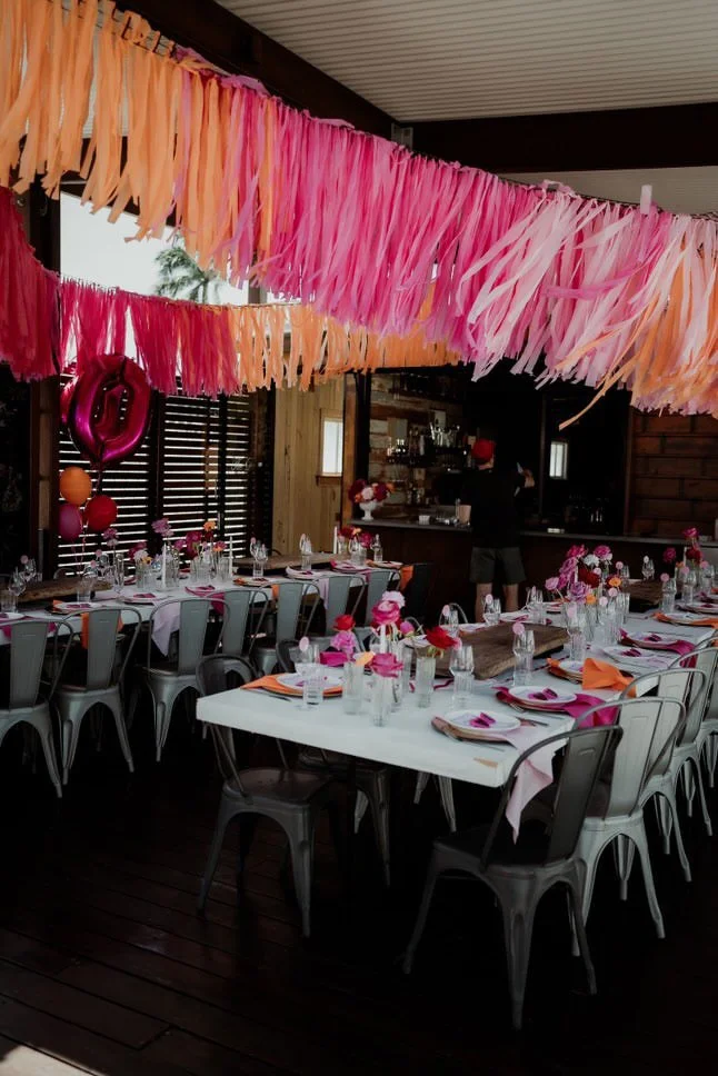 Decorated indoor dining area with pink, peach, and orange streamers hanging from ceiling, pink balloons, and tables set for a celebration or party.
