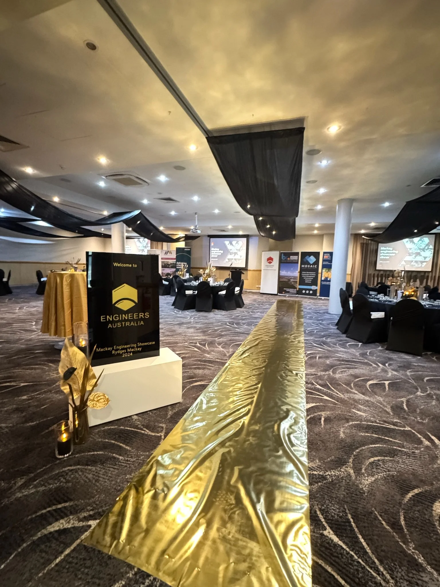 Event space decorated with black and gold accents, set for a formal gathering with tables and chairs, large screens, and banners in the background. A golden aisle runner runs through the center of the room.