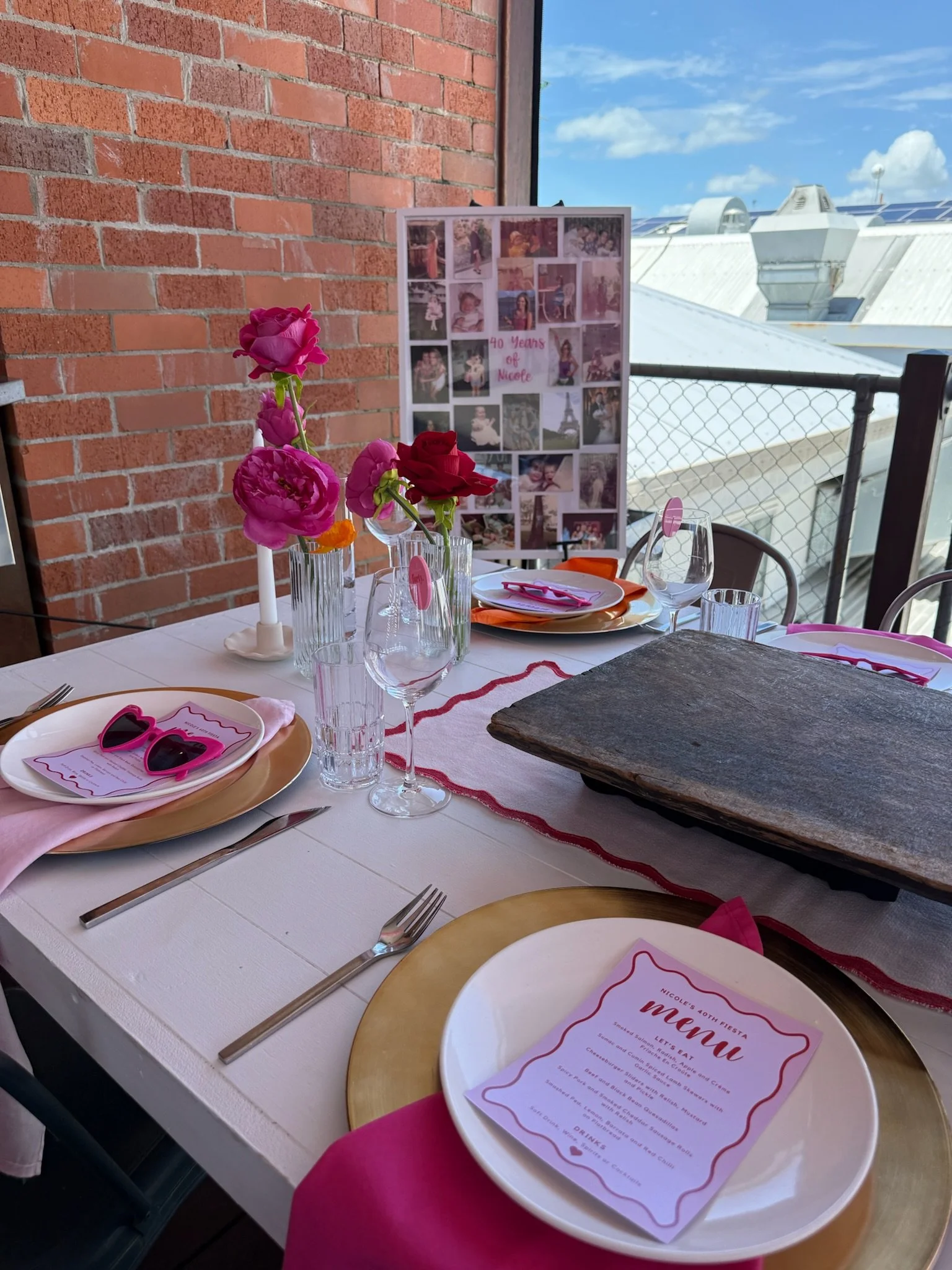 A decorated dining table with pink and red flowers in glass vases, pink sunglasses, colorful plates, and a menu for a 40th birthday celebration, with a backdrop of a brick wall and city rooftop view.