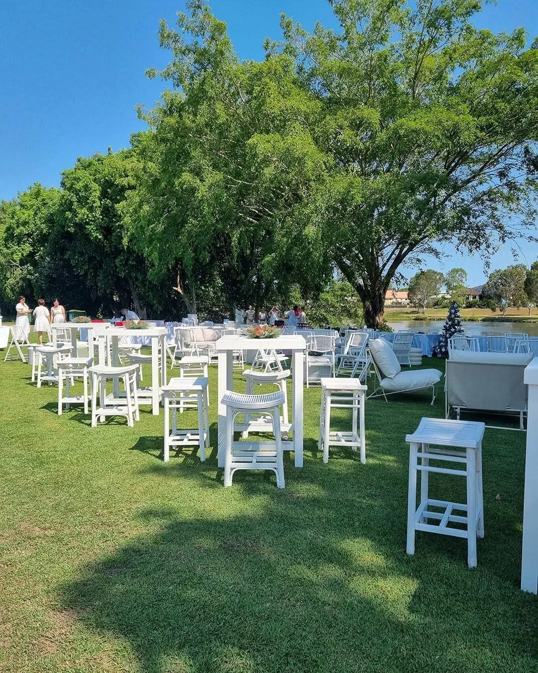 Outdoor event setup with white tables, chairs, and lounge furniture on a grassy area with large trees nearby and a lake in the background, under a clear blue sky.
