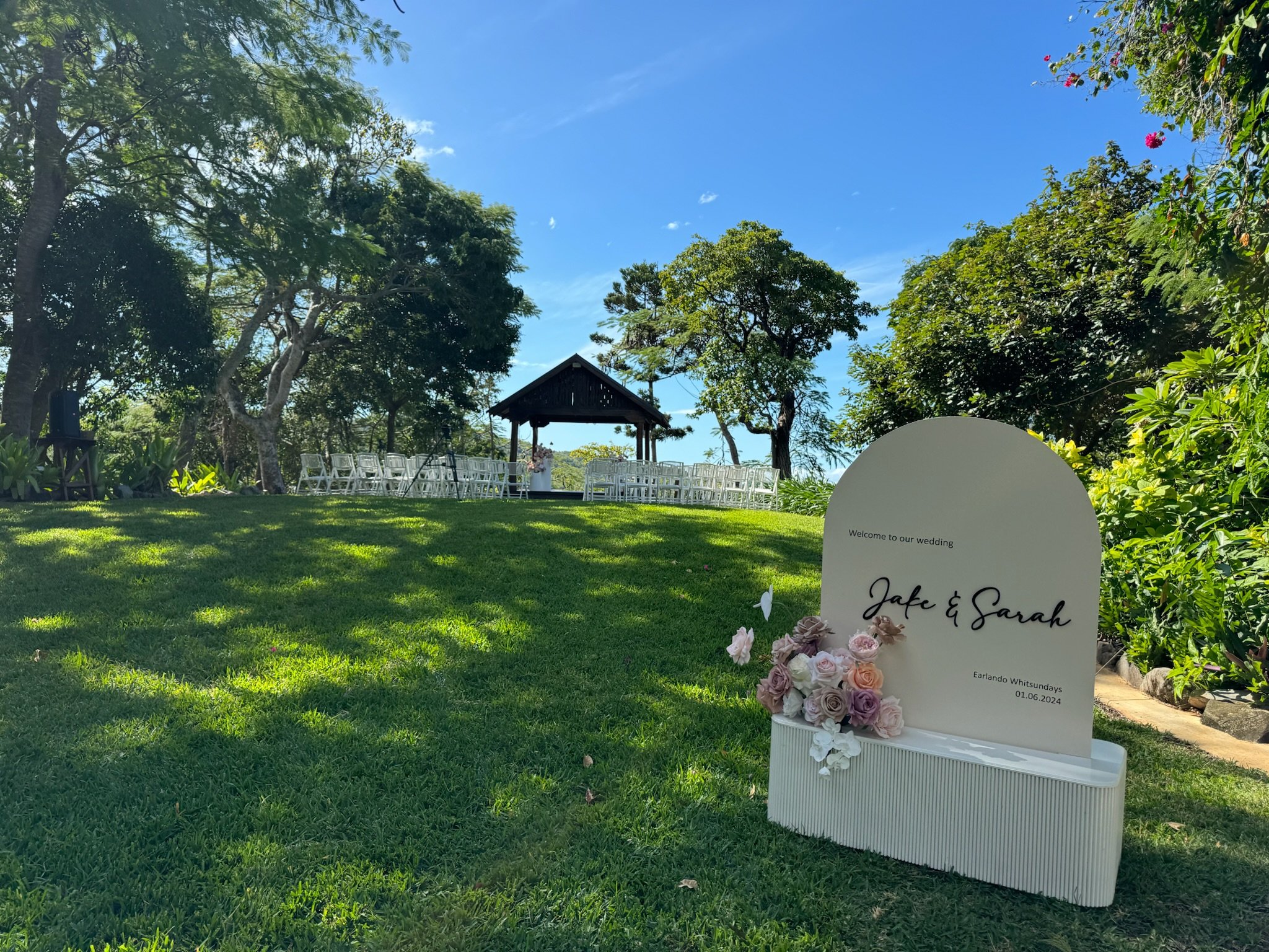 Wedding ceremony setup outdoors with white chairs, a small stage, and a welcome sign for Jake and Sarah, located on a grassy area surrounded by trees under a blue sky.