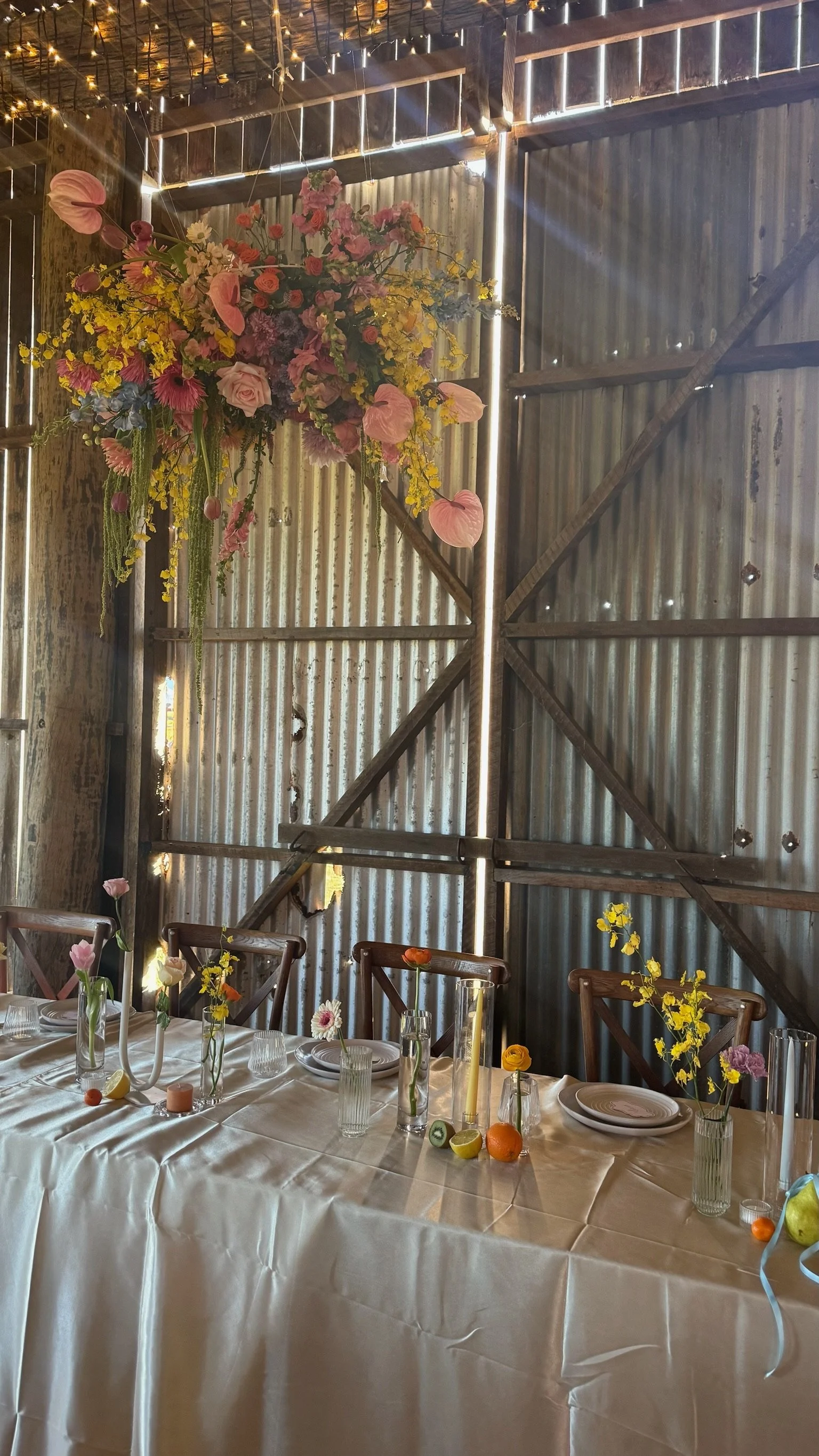 A rustic table setting inside a barn with a large floral arrangement hanging from the ceiling. The table is decorated with small vases of flowers, fruit, candles, and place settings, featuring a cream-colored tablecloth.