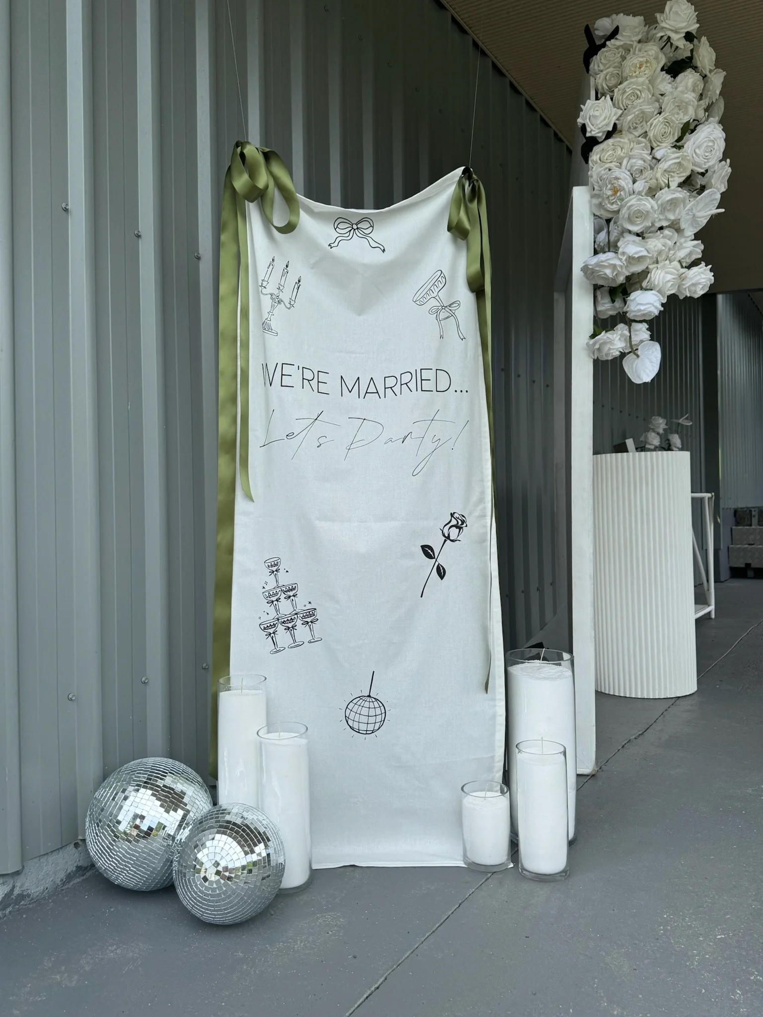 Wedding celebration display with a tall white banner that says 'We're Married... Let's Party!' Decorated with illustrations of candles, a bow, a comb, a rose, a disco ball, and champagne glasses. Several white candles in glass holders and two silver 