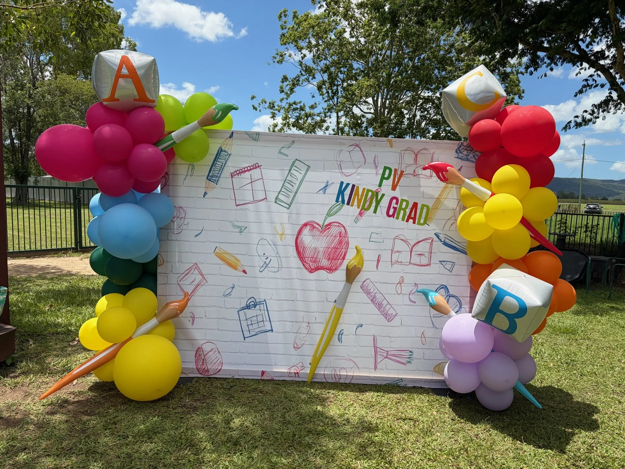 Colorful balloon arch with blocks, paintbrushes, and school-themed decorations surrounding a white brick backdrop with drawings and text saying "PV KNDY GRAD."