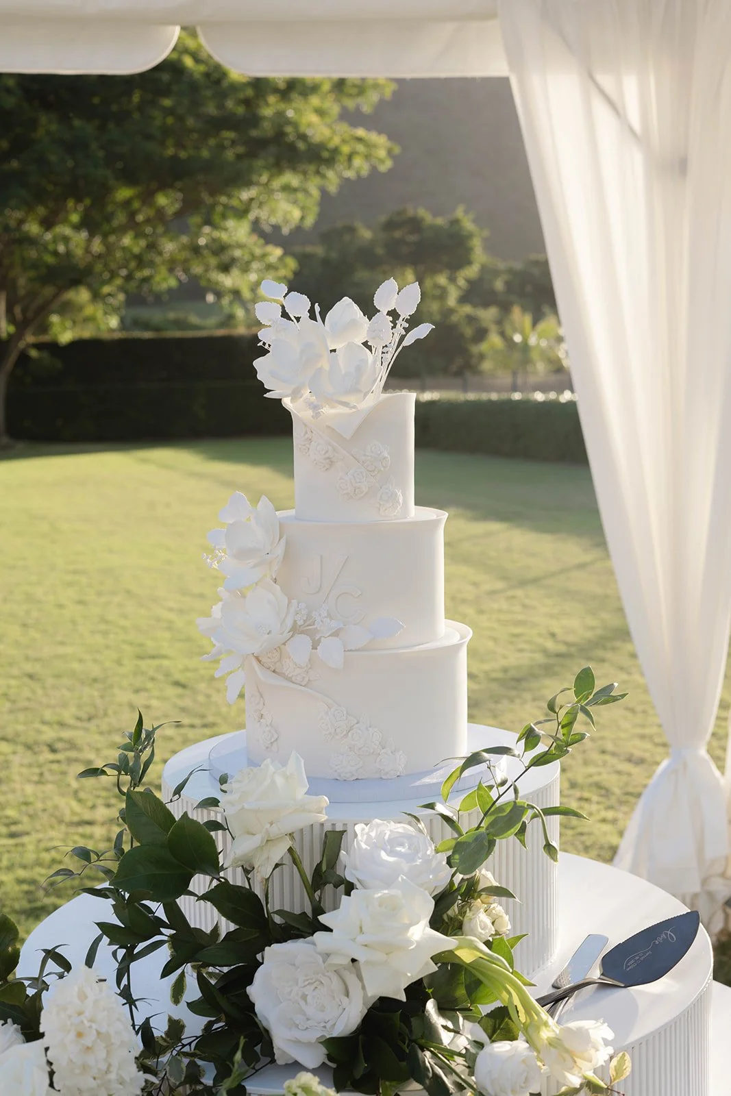 A three-tier white wedding cake decorated with white flowers and greenery, placed on a table outdoors with a sunny green landscape and white drapery in the background.