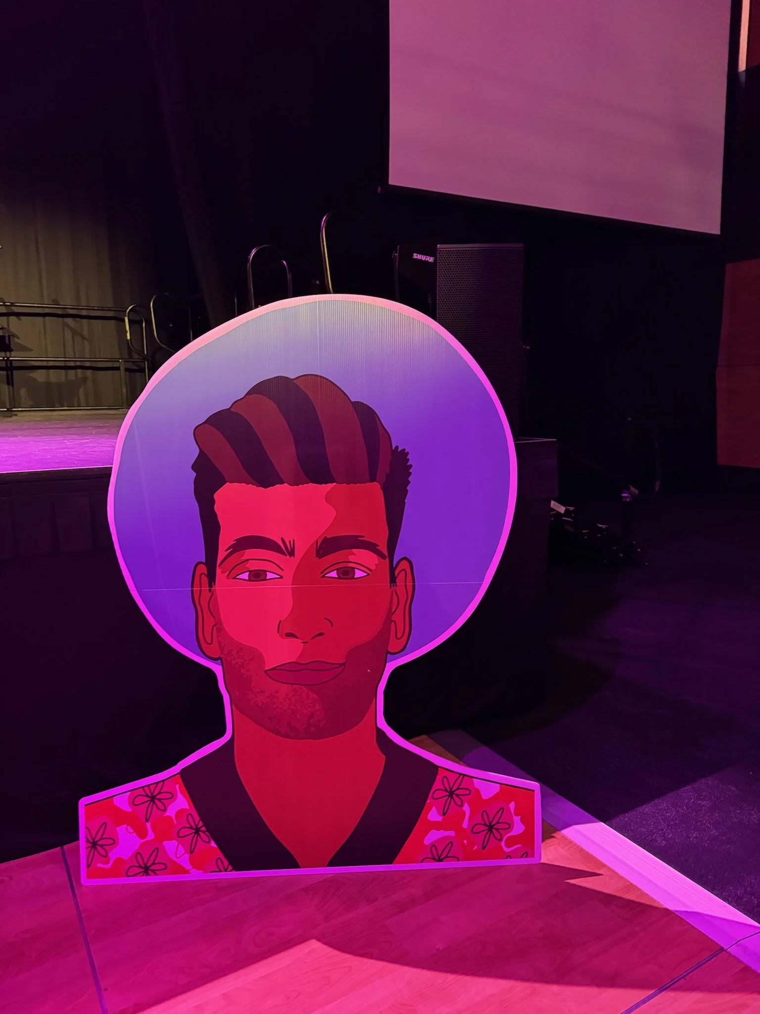 Colorful illustration of a man with a pompadour hairstyle and a slight beard, wearing a patterned shirt with flowers, illuminated by pink and purple lighting.