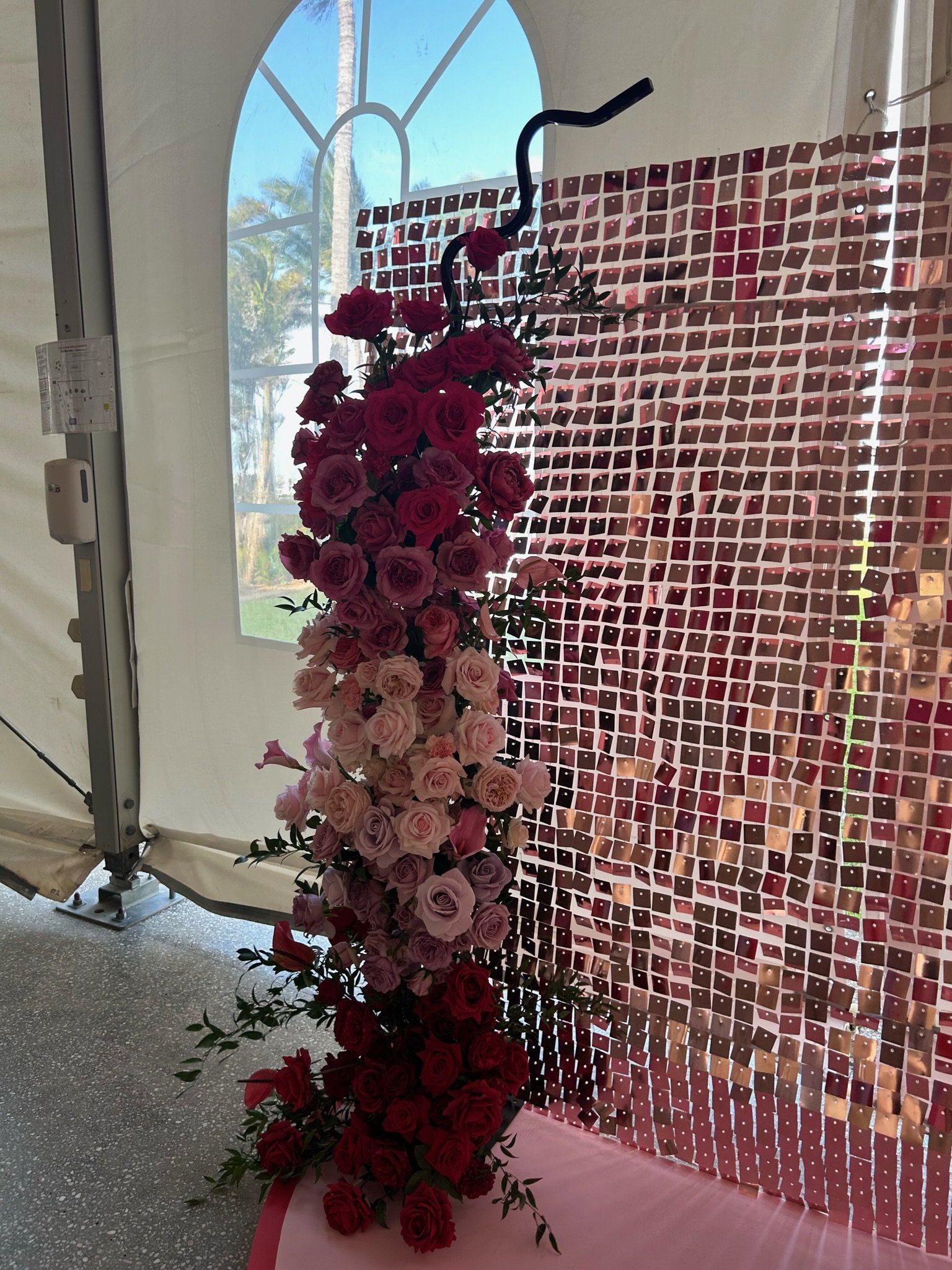 A floral arrangement of red, pink, and purple roses, cascading down, with a decorative metal sculpture of a flamingo on top, set against a backdrop of a reflective, mosaic wall and a window with a view of palm trees.
