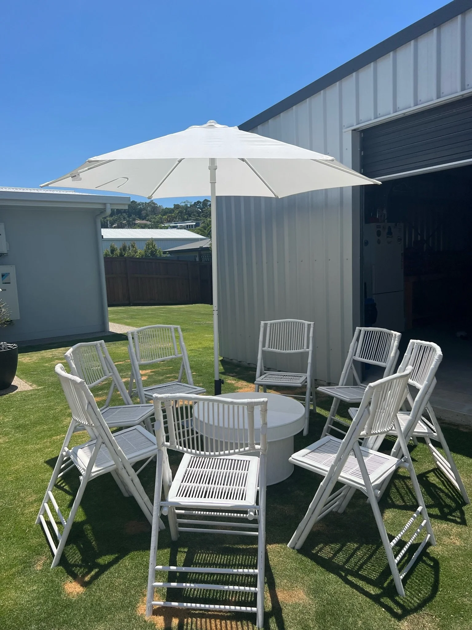 Outdoor patio area with a white umbrella, nine white chairs, and a round white table on a grassy yard outside a building.