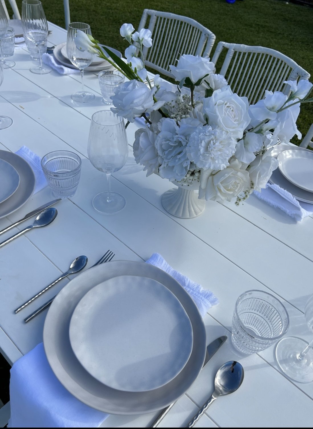 Elegant outdoor table setting with white plates, silverware, clear glasses, silk napkins, and a large white floral centerpiece with roses and other white flowers.