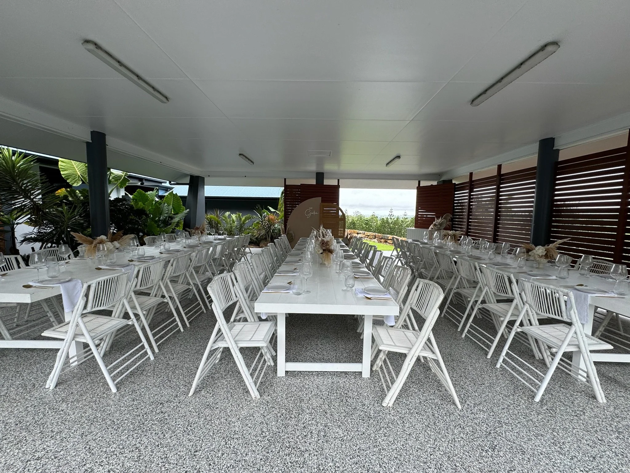 Long white banquet table set with wine glasses, plates, and napkins, surrounded by white chairs, under a covered outdoor area with plants and wooden slats on one side.