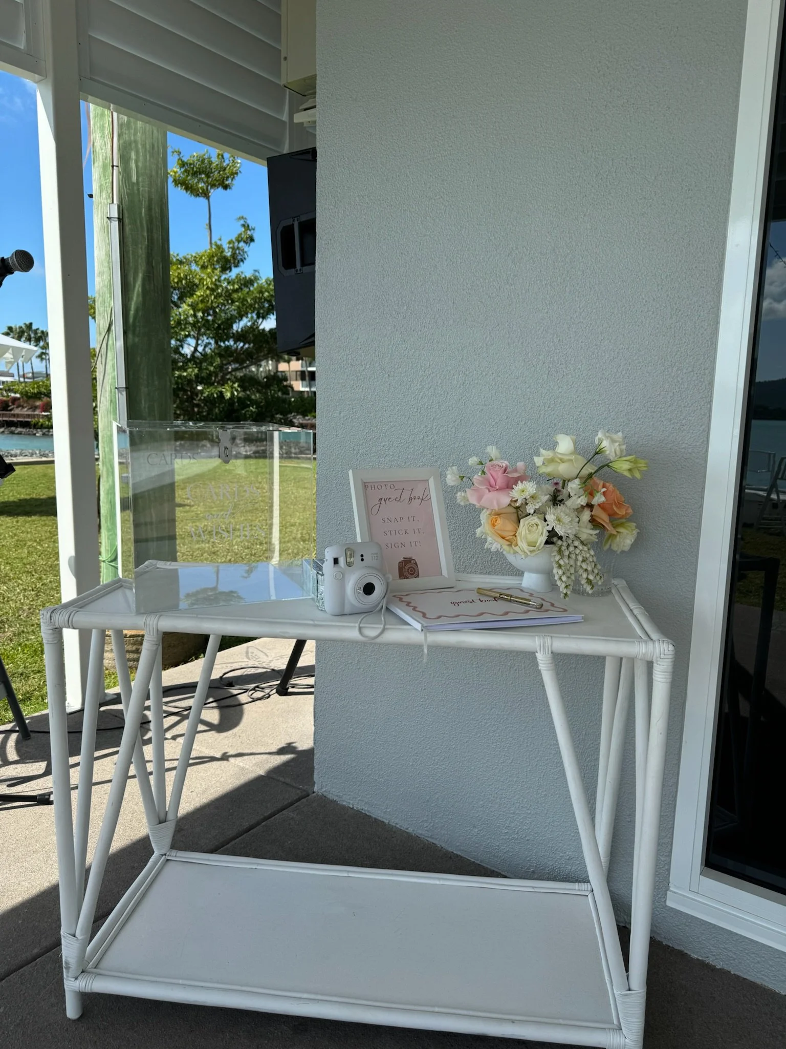 White table with flowers, signs, camera, and pen, set up for a photo booth outside next to a white wall, with a scenic outdoor background.