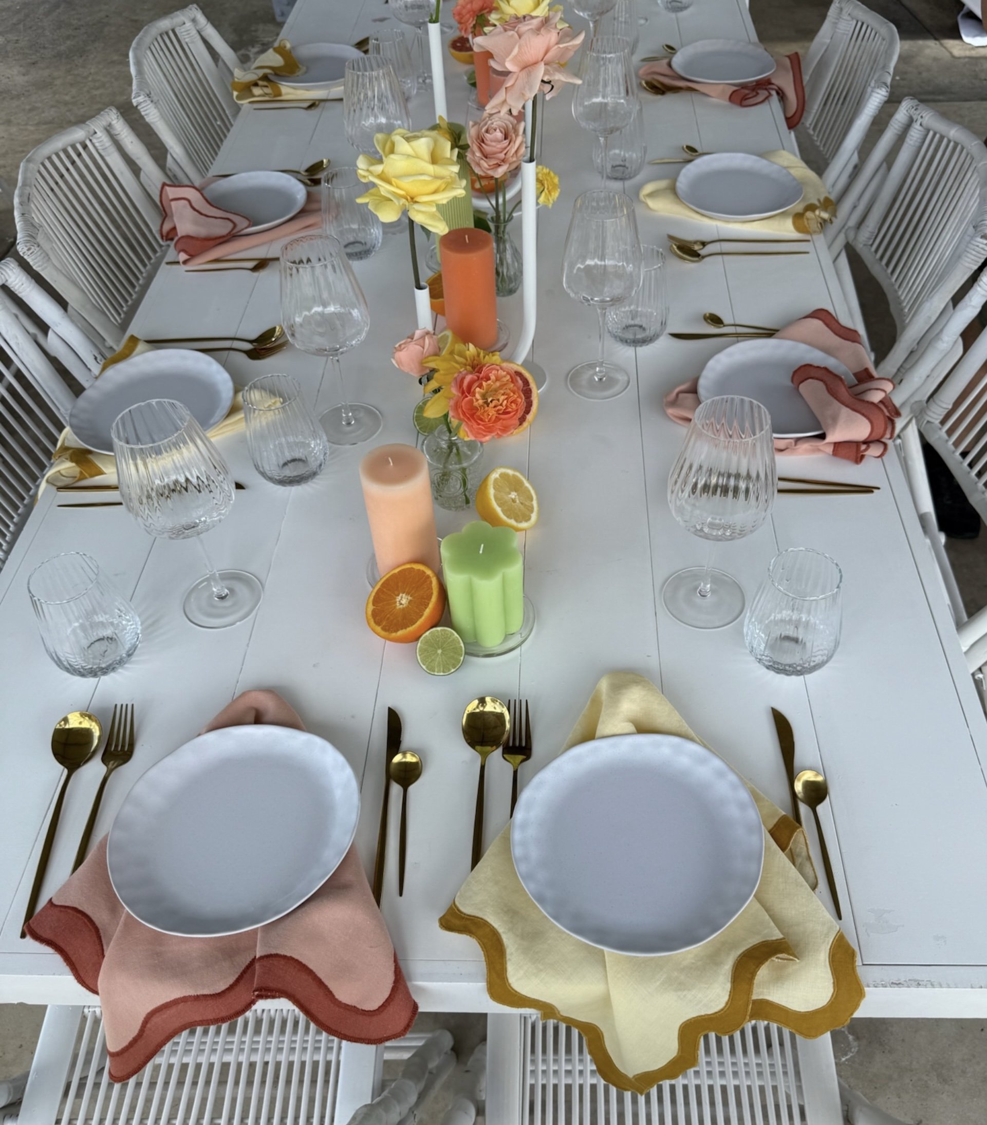A decorated dining table with white plates, gold cutlery, pink and yellow napkins, clear glassware, colorful flowers, and candles, set for a meal.