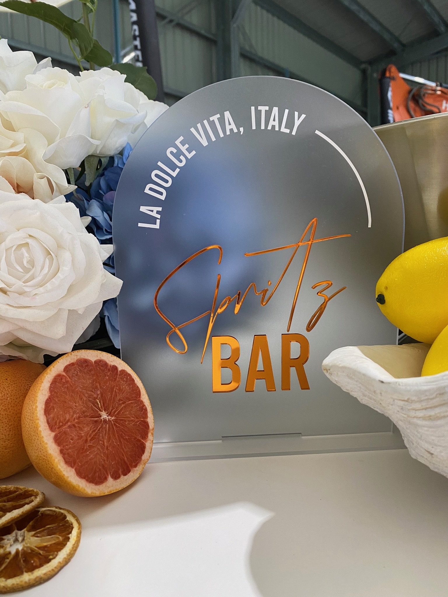 Sign with the text 'La Dolce Vita, Italy' and 'Gorizia Bar', surrounded by white roses, a halved grapefruit, and dried orange slices, in a setting resembling a bar or café.