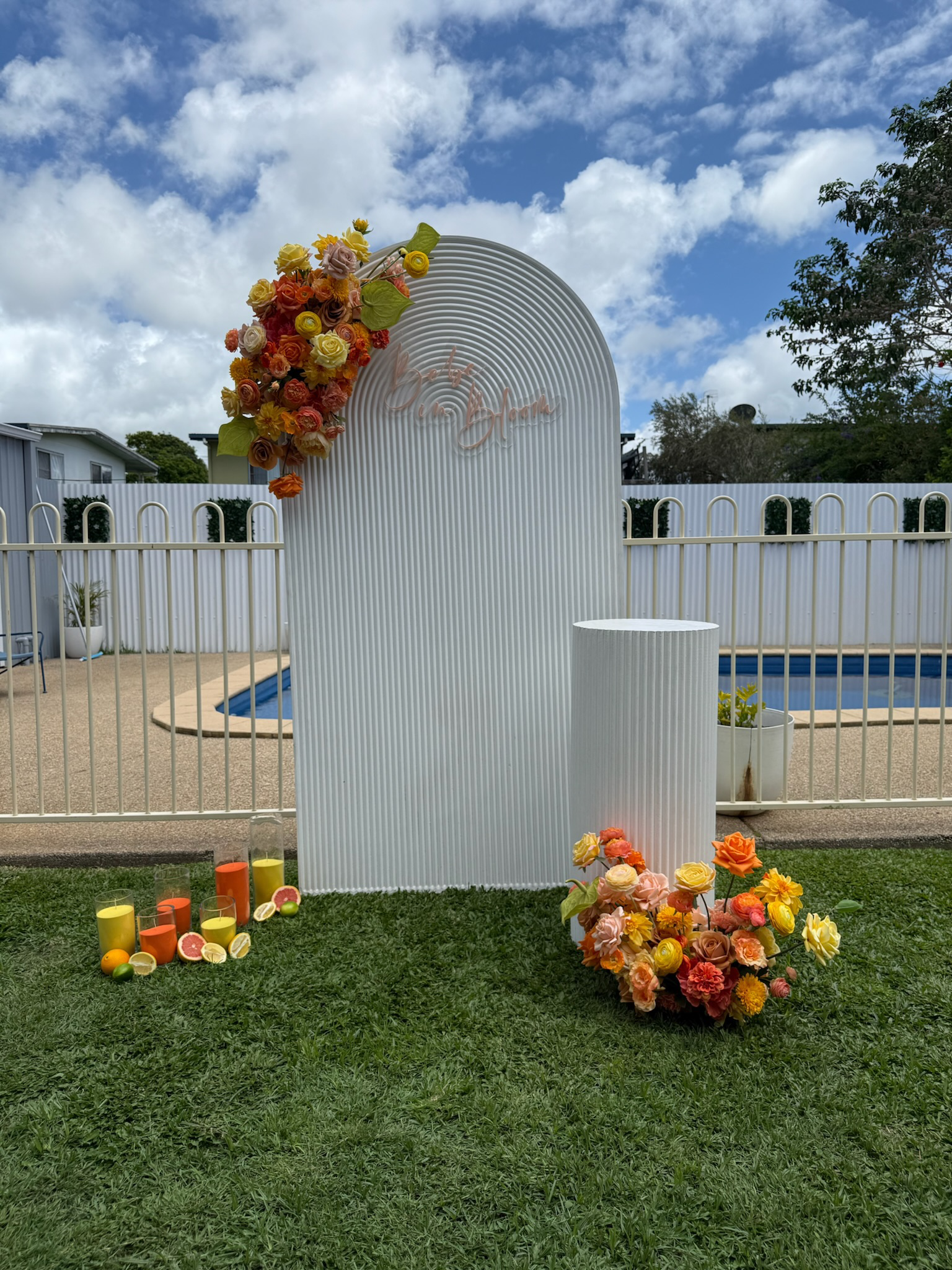 Decorative outdoor scene with a white arched backdrop decorated with pink and yellow flowers, adjacent to a white cylindrical stand, by a pool area with a white fence, colorful candles and flowers on the grass, and a partly cloudy sky overhead.