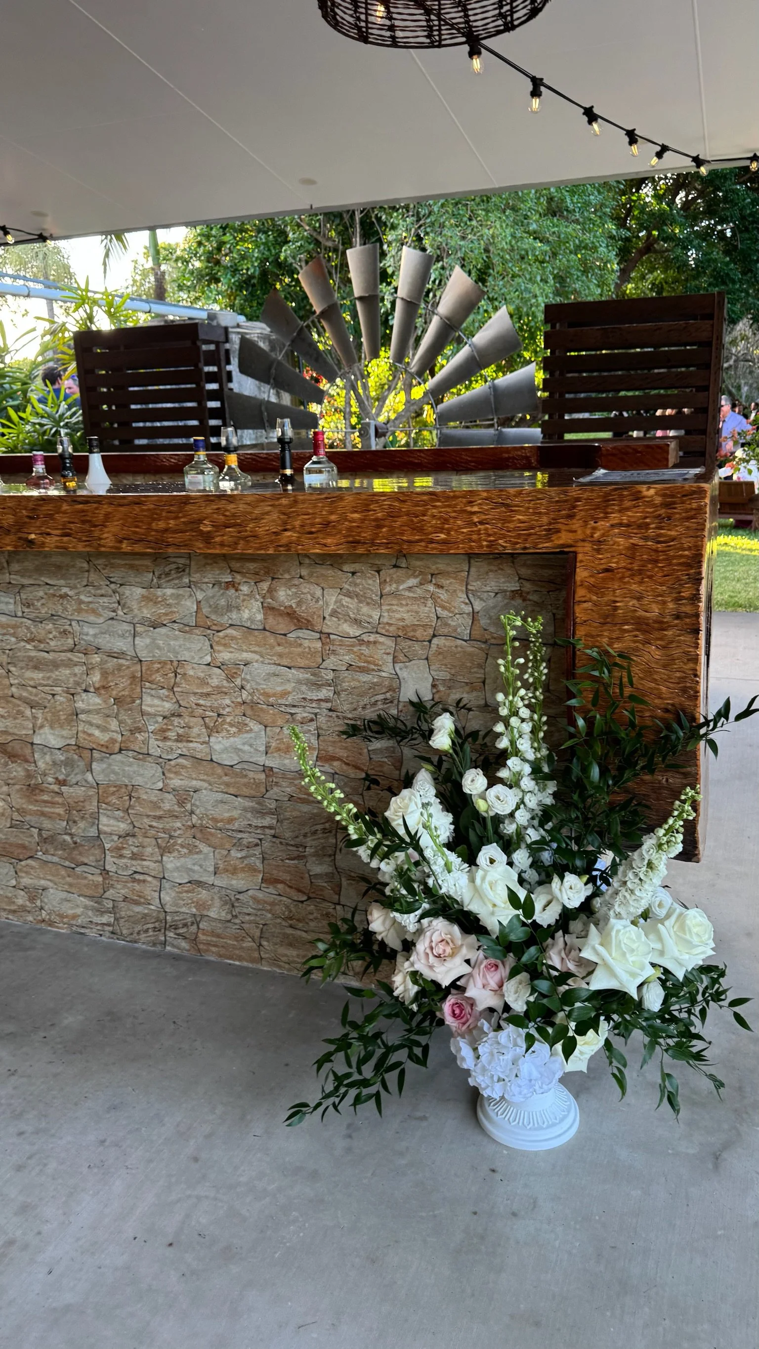 A floral arrangement with white and light pink flowers in a white vase, placed in front of a stone and wood counter, outdoors.