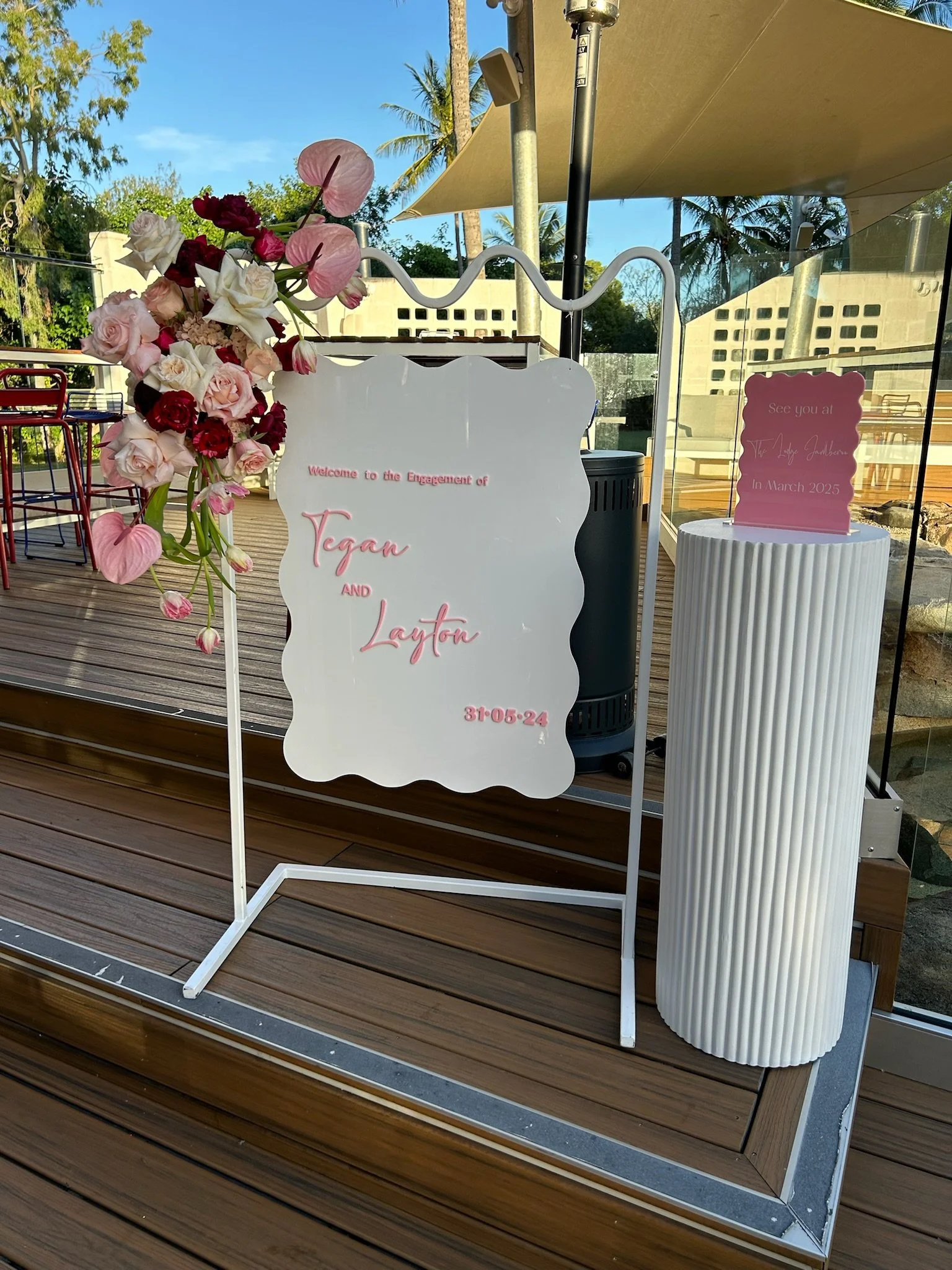 Wedding reception sign with floral decoration, welcoming Tegan and Layton, displaying the time 31:05:24, and a smaller sign indicating the couple's wedding date in March 2025, set outdoors on a wooden deck.