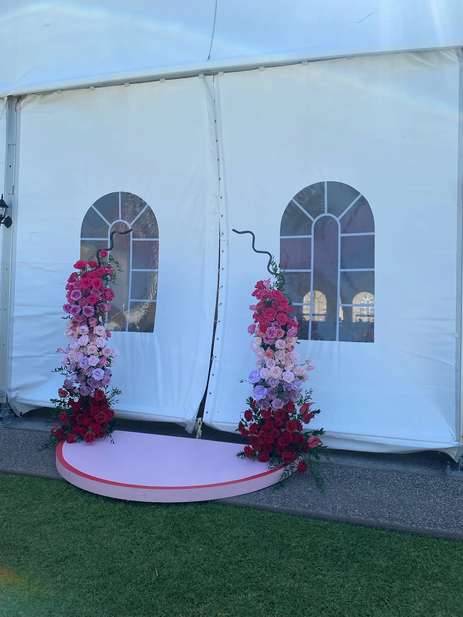 Decorative wedding arch with pink, purple, and red roses, set against white tent walls with window graphics.