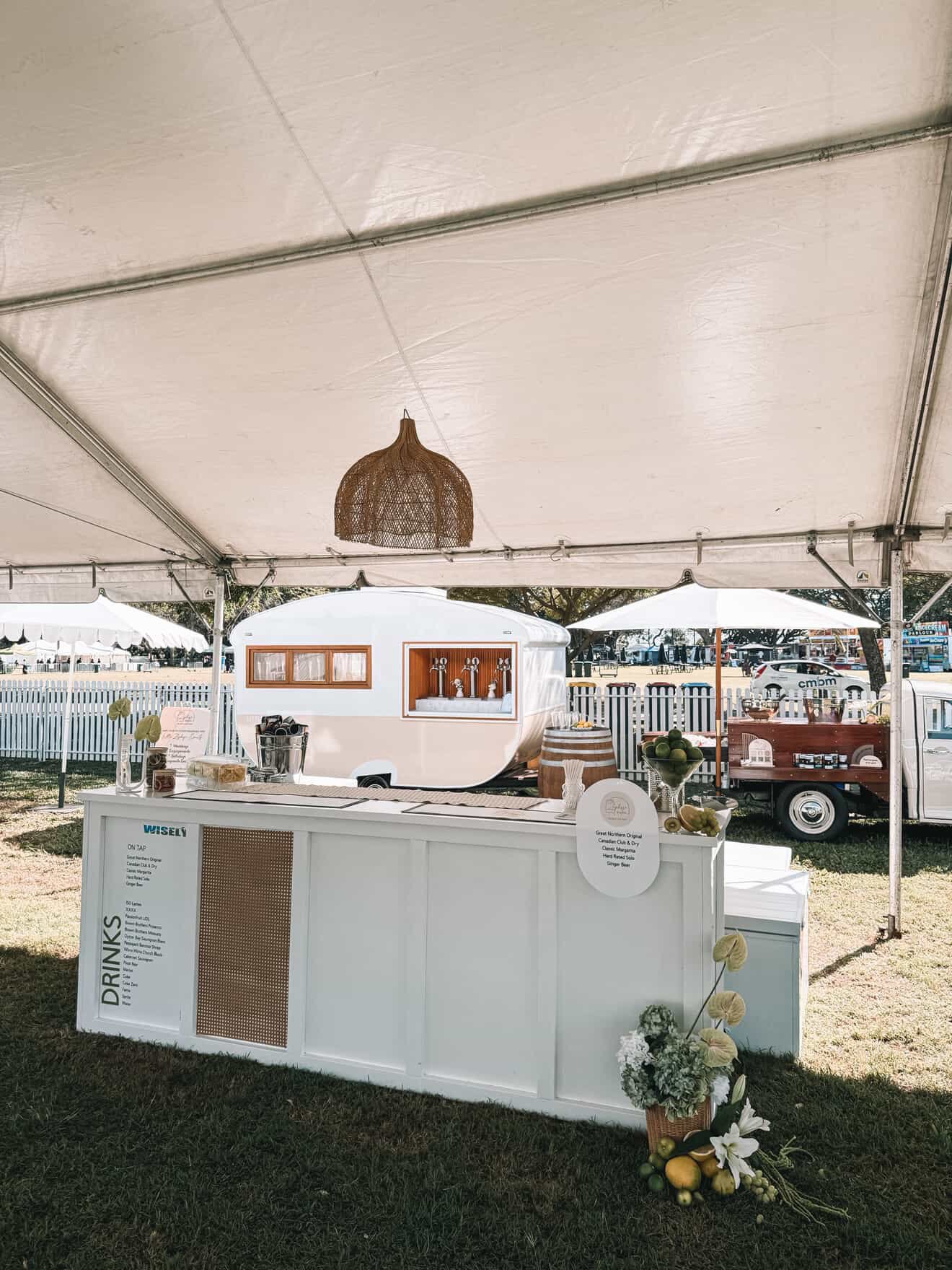 A white vintage trailer set up as a beverage stand at an outdoor event, decorated with flowers and a wooden bar, with a white fence and trees in the background.