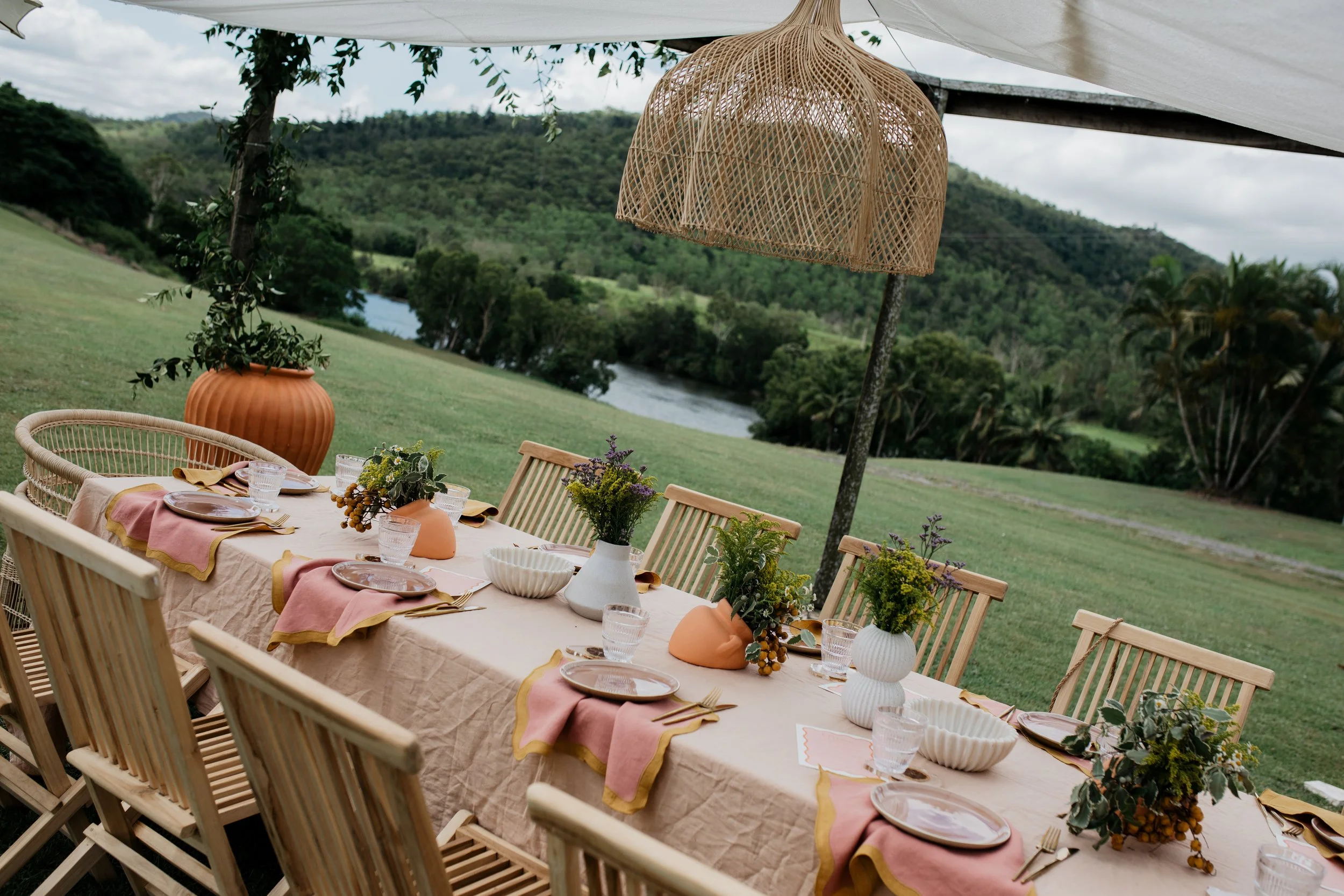 Outdoor dining table set with pink and yellow cloth napkins, plates, glasses, cutlery, and floral arrangements in vases, under a woven rattan light fixture with a scenic view of a river, green hills, and trees in the background.
