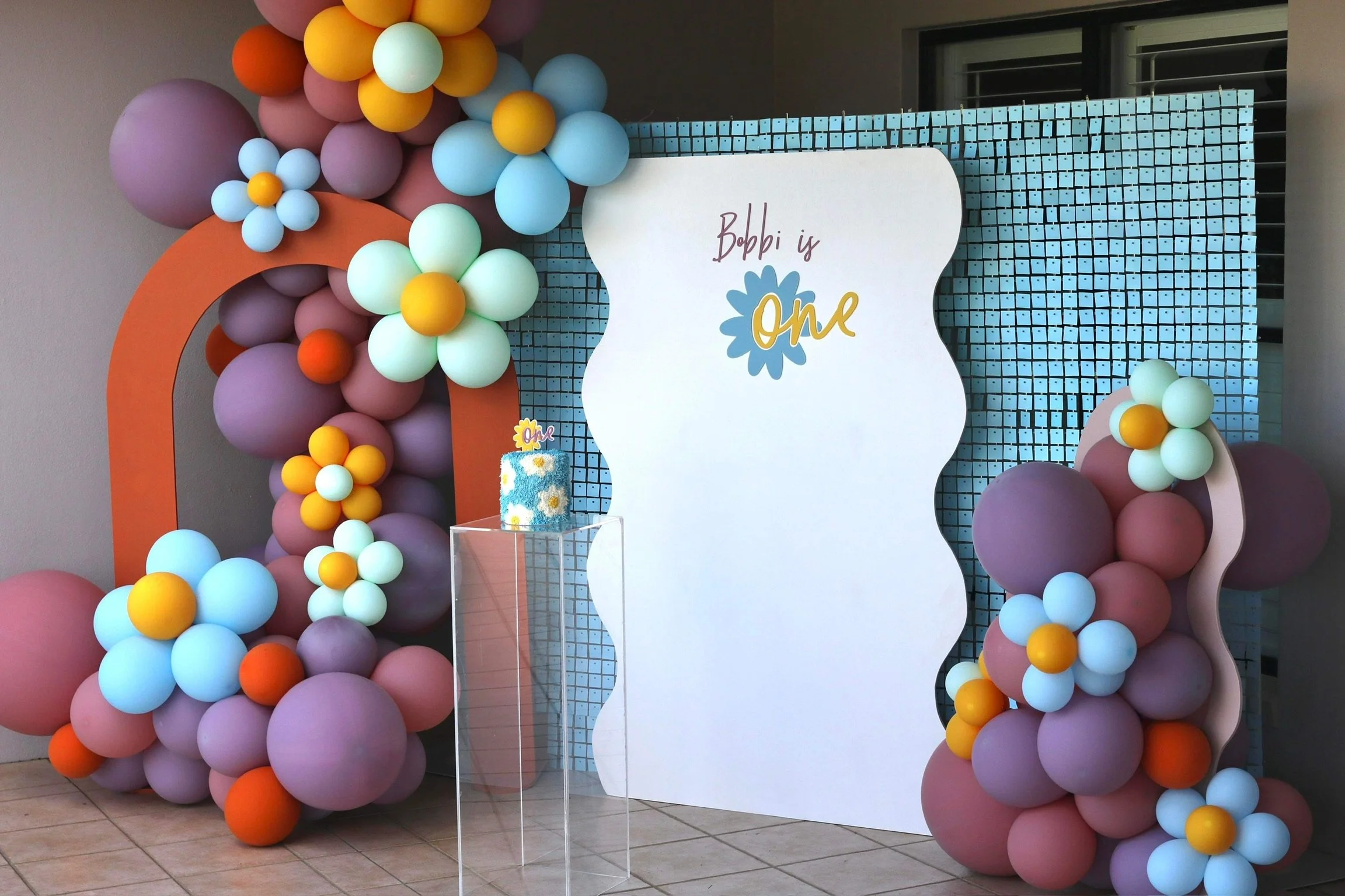 Decorative backdrop with a blue grid pattern, large white sign that reads 'Bobby is One' with a blue flower, colorful balloon arrangements with pastel and bright colors, a small birthday cake with daisy decorations and a 'One' topper.
