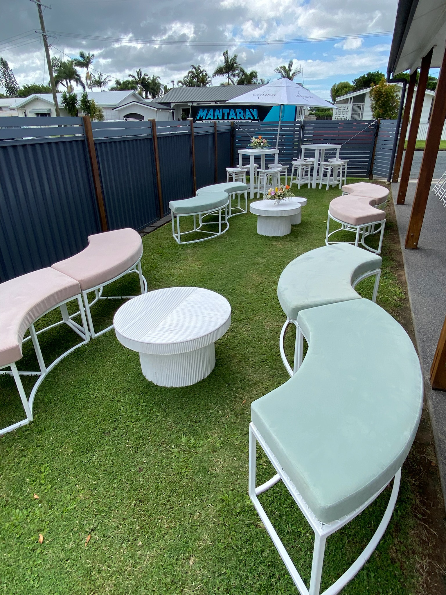 Outdoor seating area with pastel-colored curved benches, round tables, and flower arrangements on a grass lawn. There is a blue fence, a large umbrella, and residential houses with palm trees in the background under a partly cloudy sky.