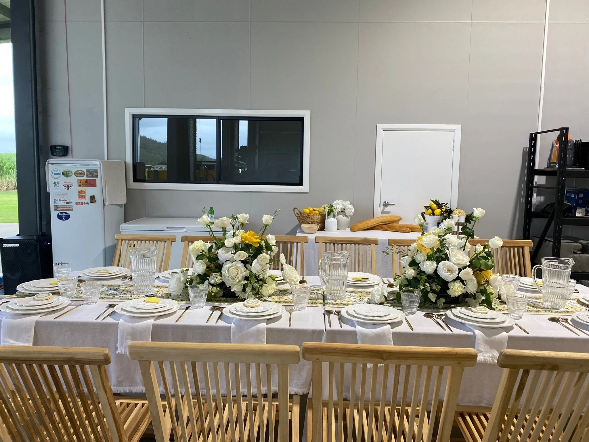 Dining table set for a meal with white plates, silverware, and glassware, decorated with white and yellow floral centerpieces, in a room with a window, refrigerator, and shelving.