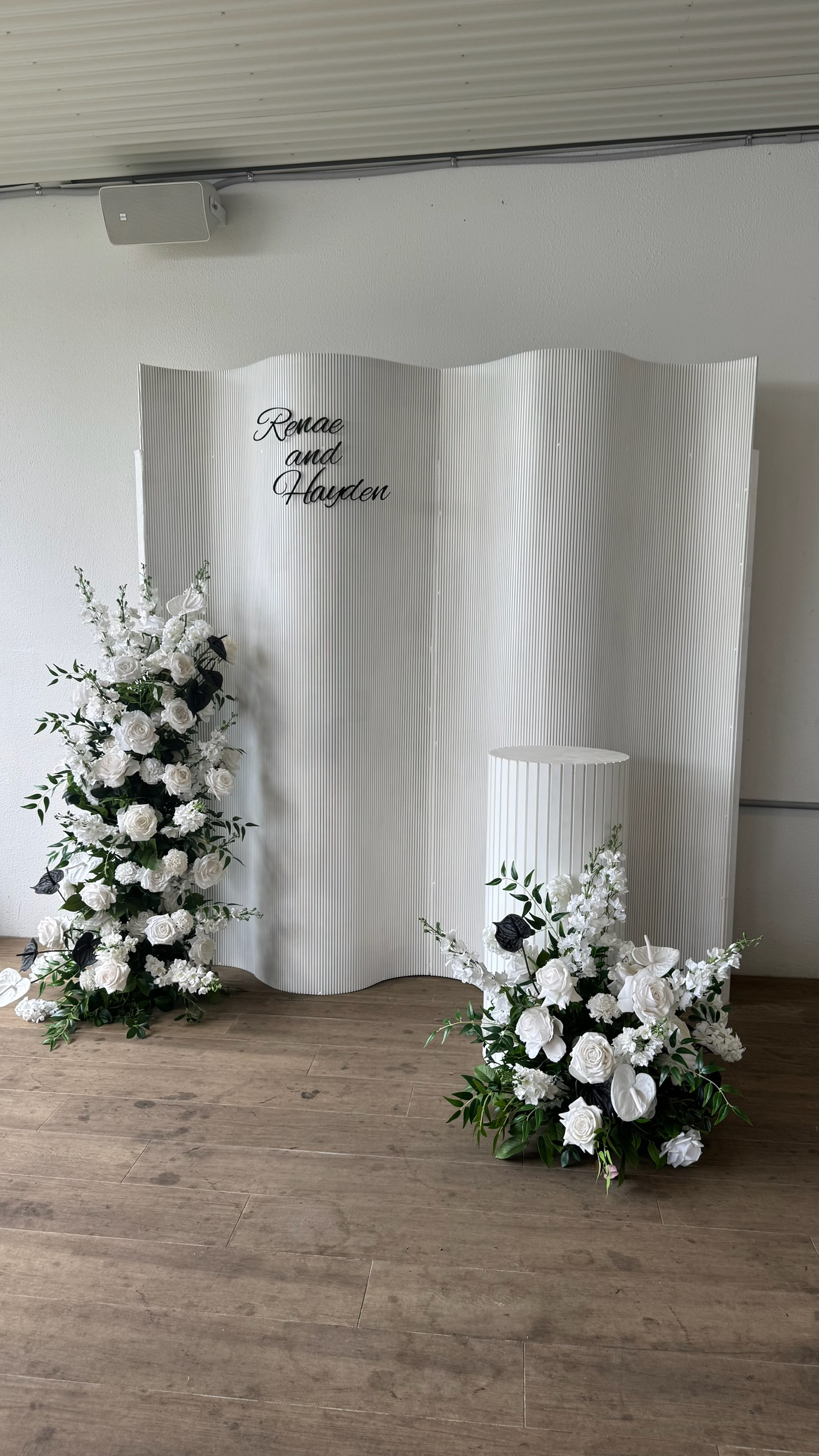 White floral arrangement and a white decorative stand in front of a wedding backdrop with the names 'Renee and Hayden' written in black cursive.