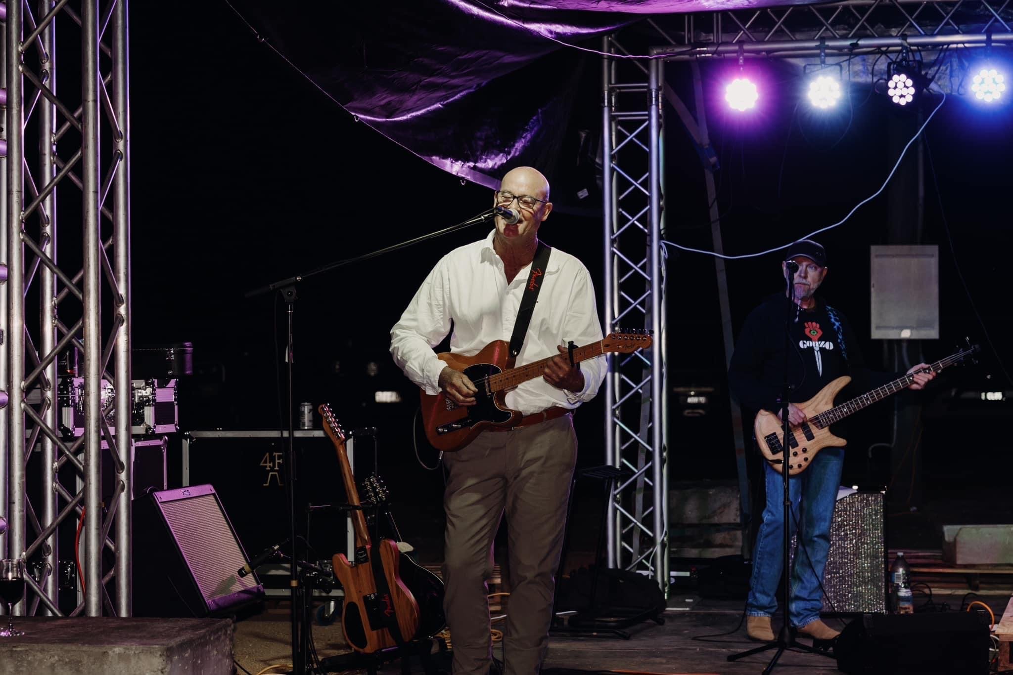Musicians performing on a stage at night, with one playing an electric guitar and singing into a microphone, and another playing a bass guitar, under purple stage lights.