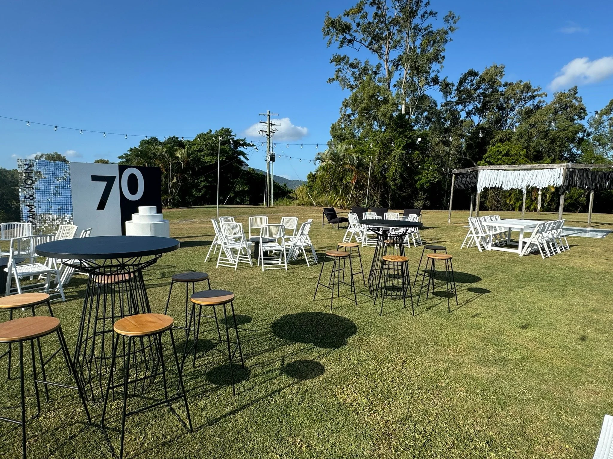 An outdoor event space with tables, chairs, and string lights on a grassy area under a clear blue sky. There is a black and white sign with the number 70.