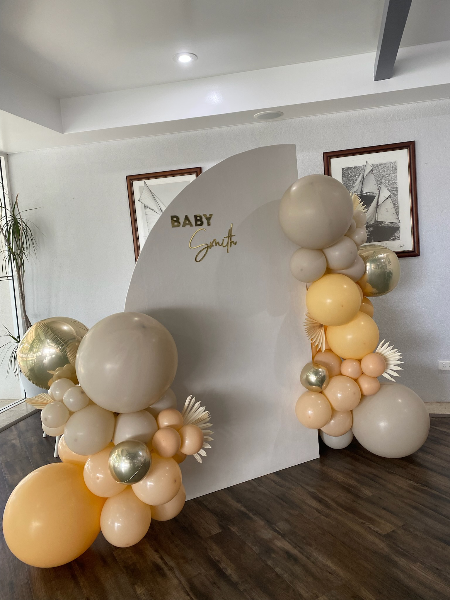 A baby gender reveal backdrop decorated with clusters of cream, gold, and peach balloons arranged around a white curved panel with the words "Baby Smith" on it. The decor includes large spherical balloons, smaller matte balloons, metallic balloons, a