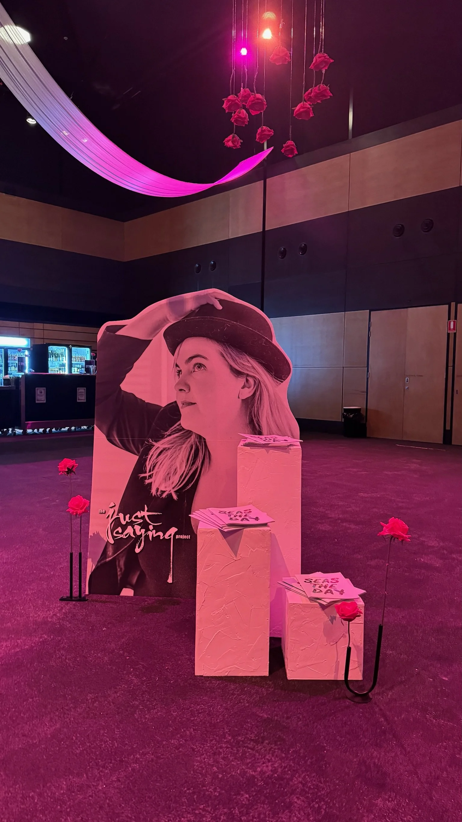 A promotional display features a large black-and-white cutout of a woman wearing a hat, with the words 'the just saying project' written on it. Surrounding the cutout are pink artificial roses on sticks and stacks of flyers or brochures on white text