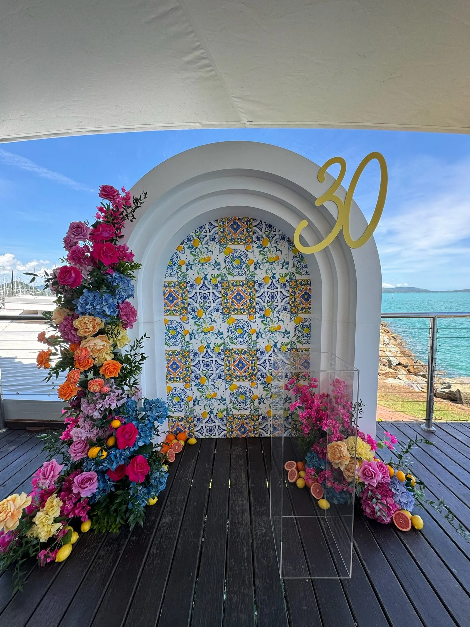 Decorated arch with colorful flowers and a yellow '30' sign, overlooking water with boats in the background, for a 30th celebration.