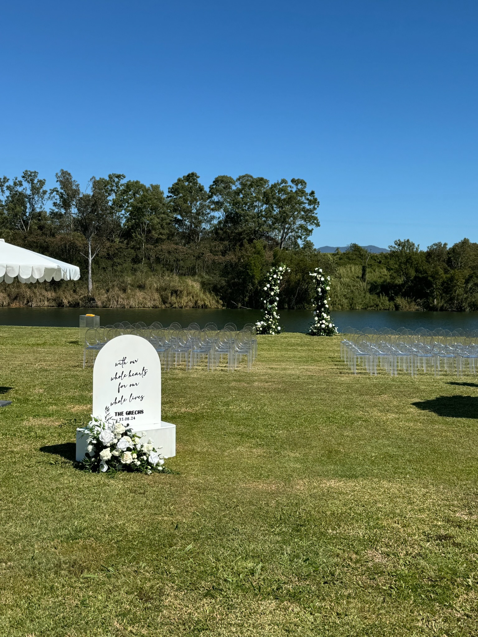 Outdoor wedding setup by a lake with white floral arch, clear chairs, a sign with white flowers, and a white tent on the left, under a blue sky with trees in the background.