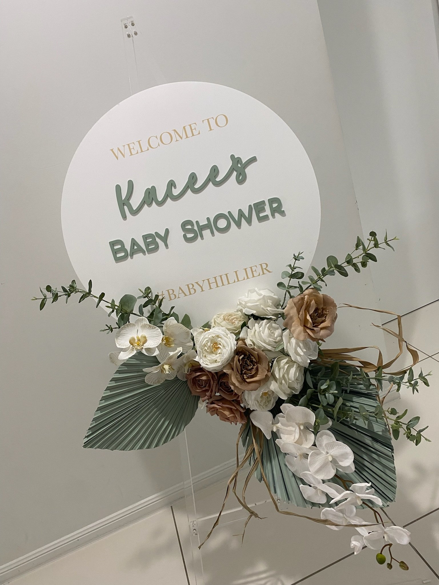 Welcome sign reading 'Welcome to Kace's Baby Shower #BABYHILLER' with a floral arrangement of white and brown flowers and greenery attached to a stand.