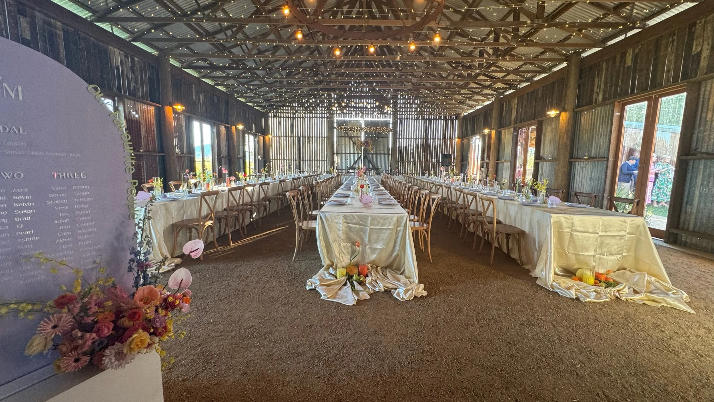 Long banquet table set inside a rustic barn with string lights, decorated with flowers in vases and tableware, preparing for a wedding reception.