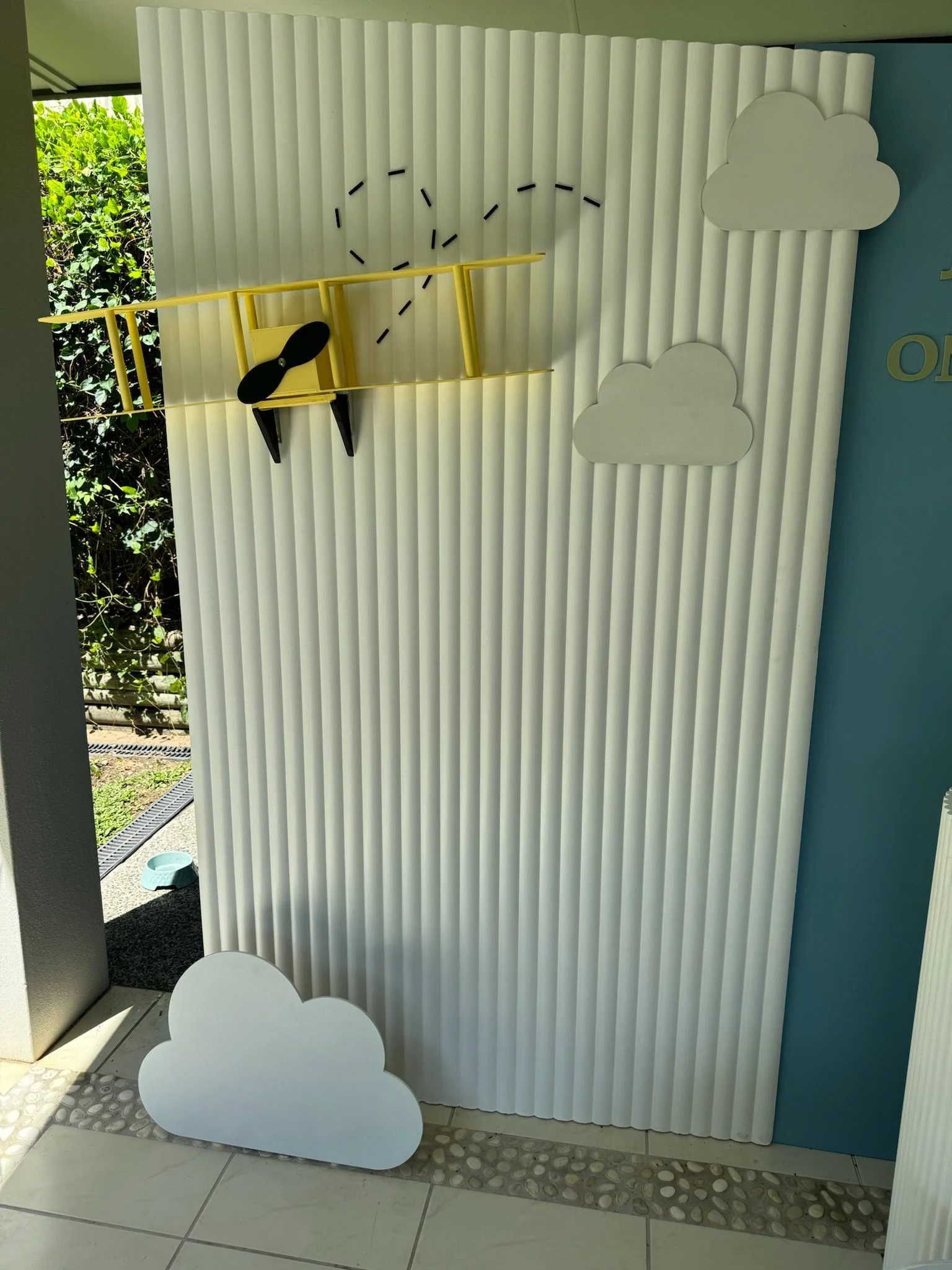 A cloud-themed wall decoration with a yellow toy airplane hanging in front, creating a shadow that looks like the airplane's shadow in the sky. The background features a white, ridged surface with cloud shapes on the right side.