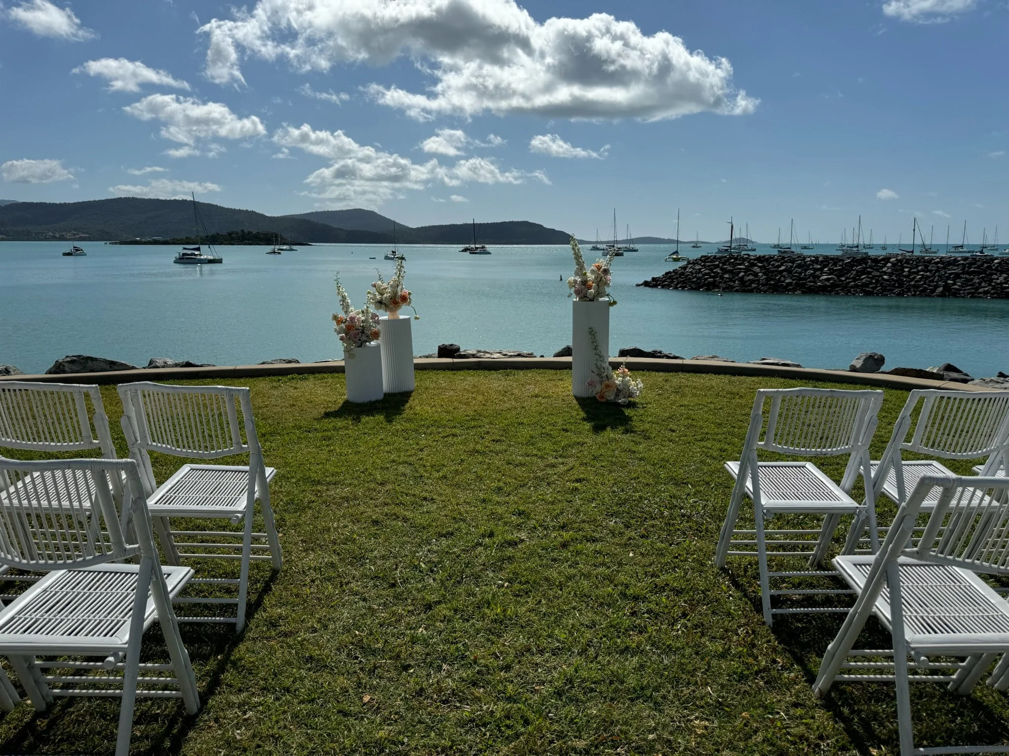 Outdoor wedding setup with white chairs facing a water backdrop, decorated with floral arrangements on tall white pedestals, boats on the water, and a cloudy sky.