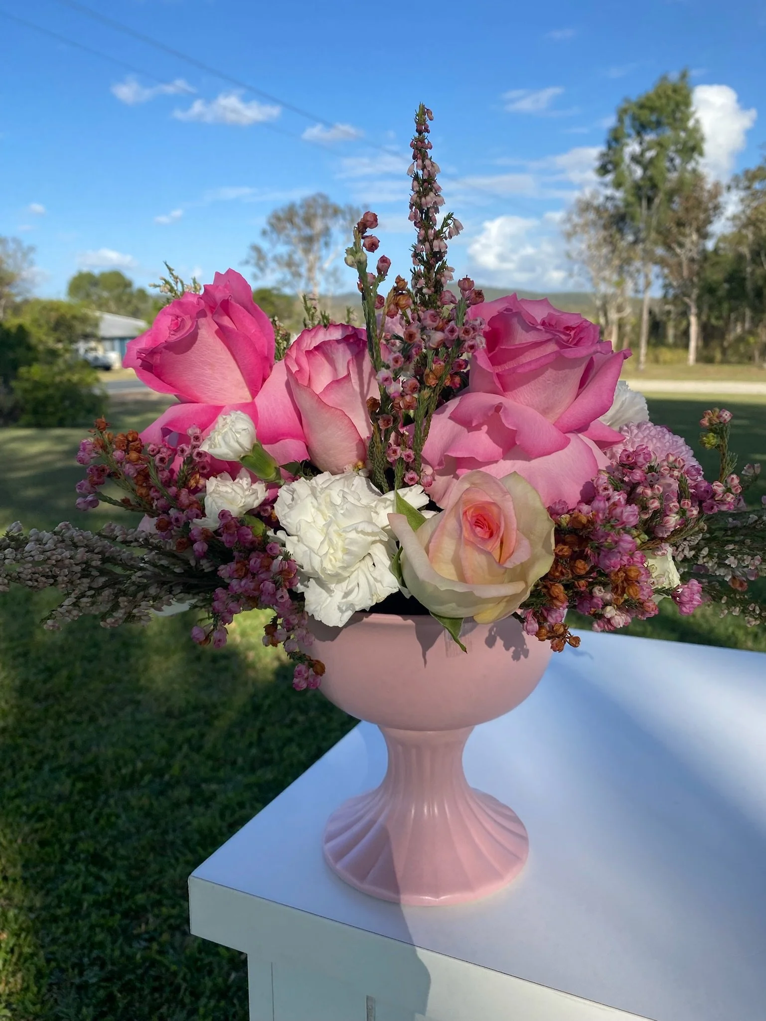A pink vase filled with pink and white roses, purple flowers, and other greenery, placed on a white surface outdoors with trees, grass, and a blue sky in the background.