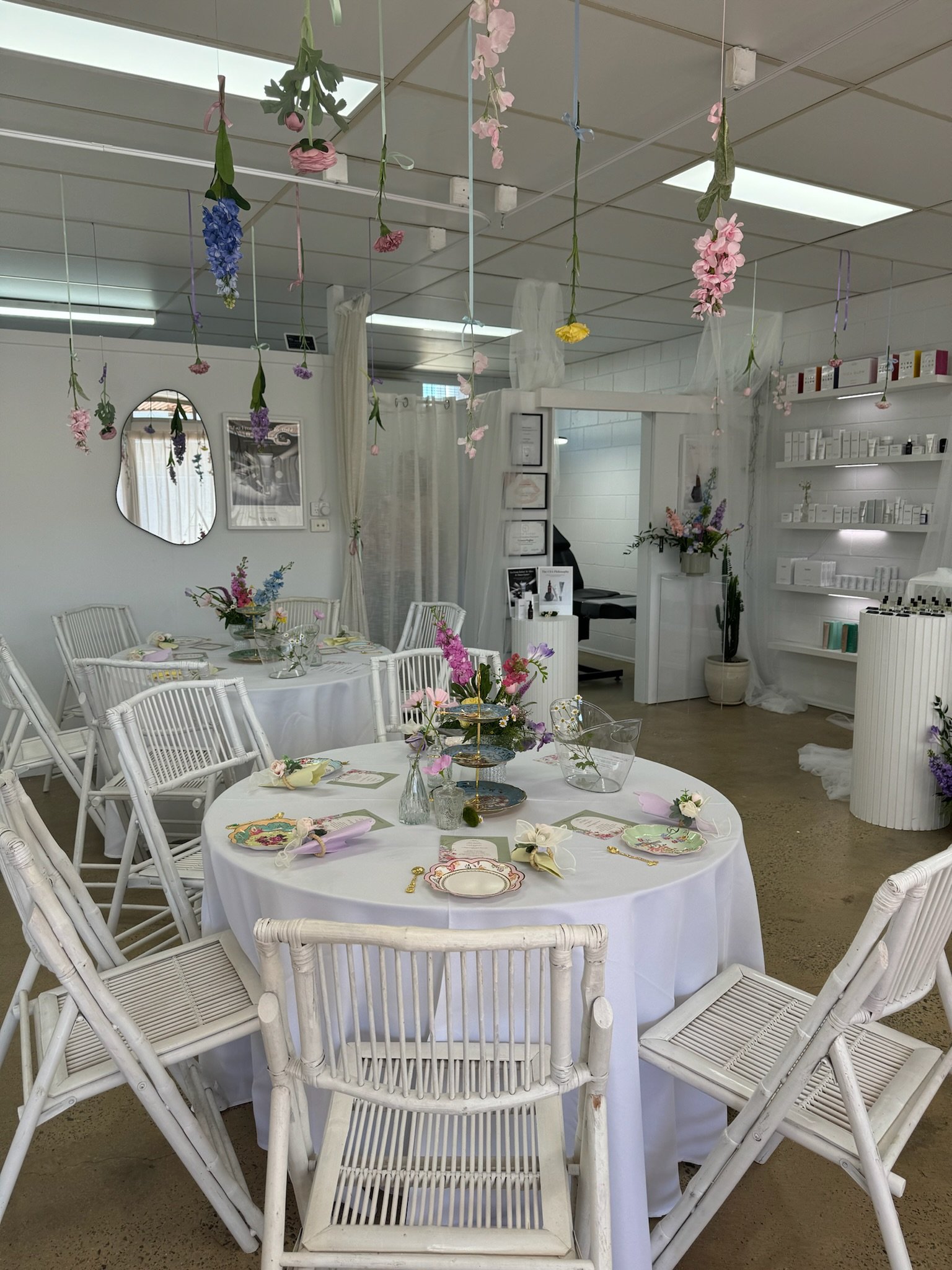 A decorated dining area with white wicker chairs and round tables covered with white tablecloths, adorned with floral centerpieces and pastel-colored plates and napkins. Hanging artificial flowers are suspended from the ceiling.