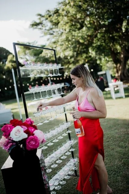 Woman in pink top and red skirt filling glasses at an outdoor champagne station with flowers in a black vase.