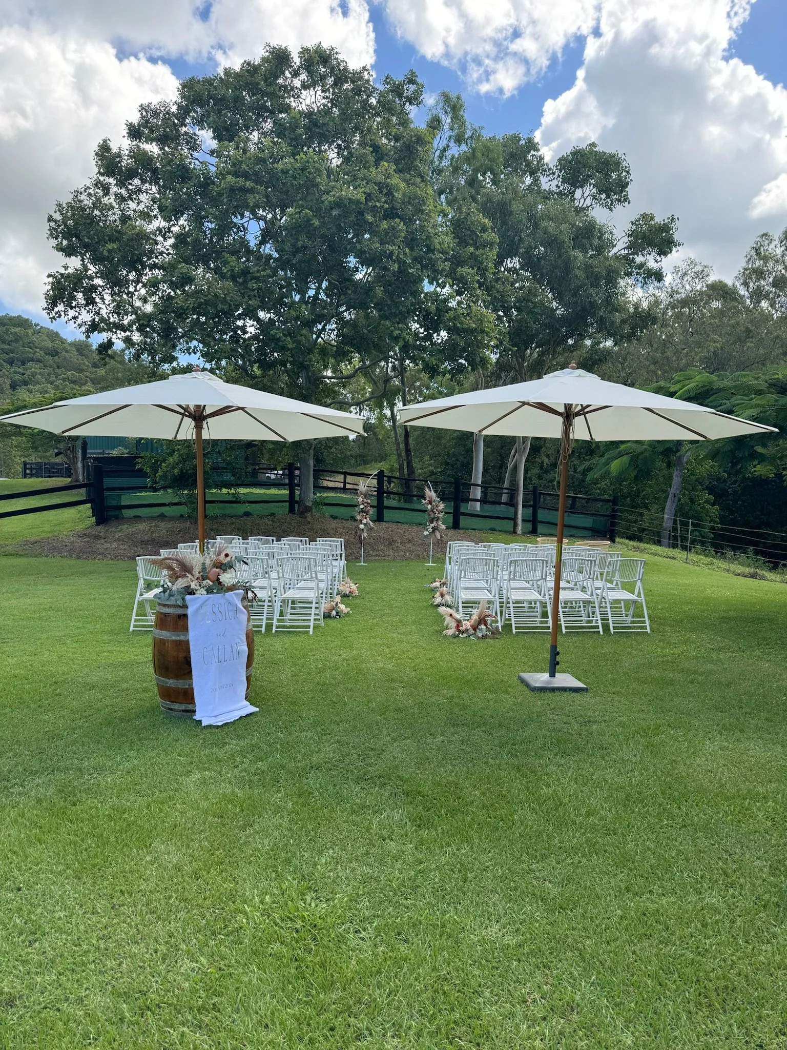 An outdoor wedding setup on a grassy area with two white umbrellas, white chairs arranged in rows, decorated with floral arrangements, a wooden barrel with a wedding sign, and beautiful trees in the background.
