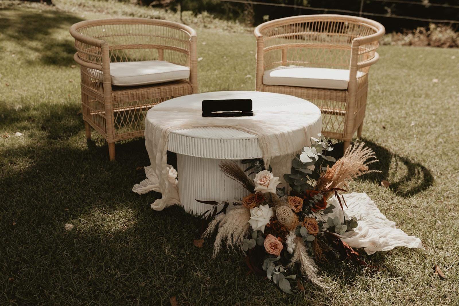 Two wicker chairs with cushions, a round table with a cloth draped over it, and a floral arrangement on the ground outside in a grassy area.