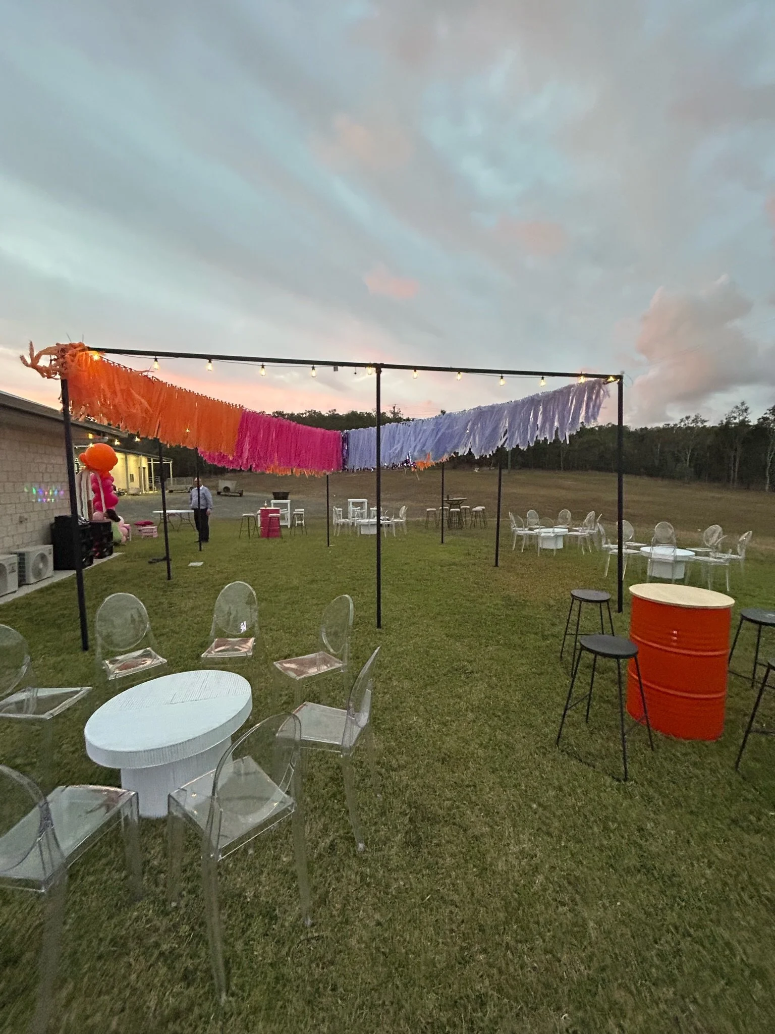 Outdoor party setup with colorful paper tassel garland, clear plastic chairs, white and black tables, orange barrel table, and string lights in a grassy area during sunset.
