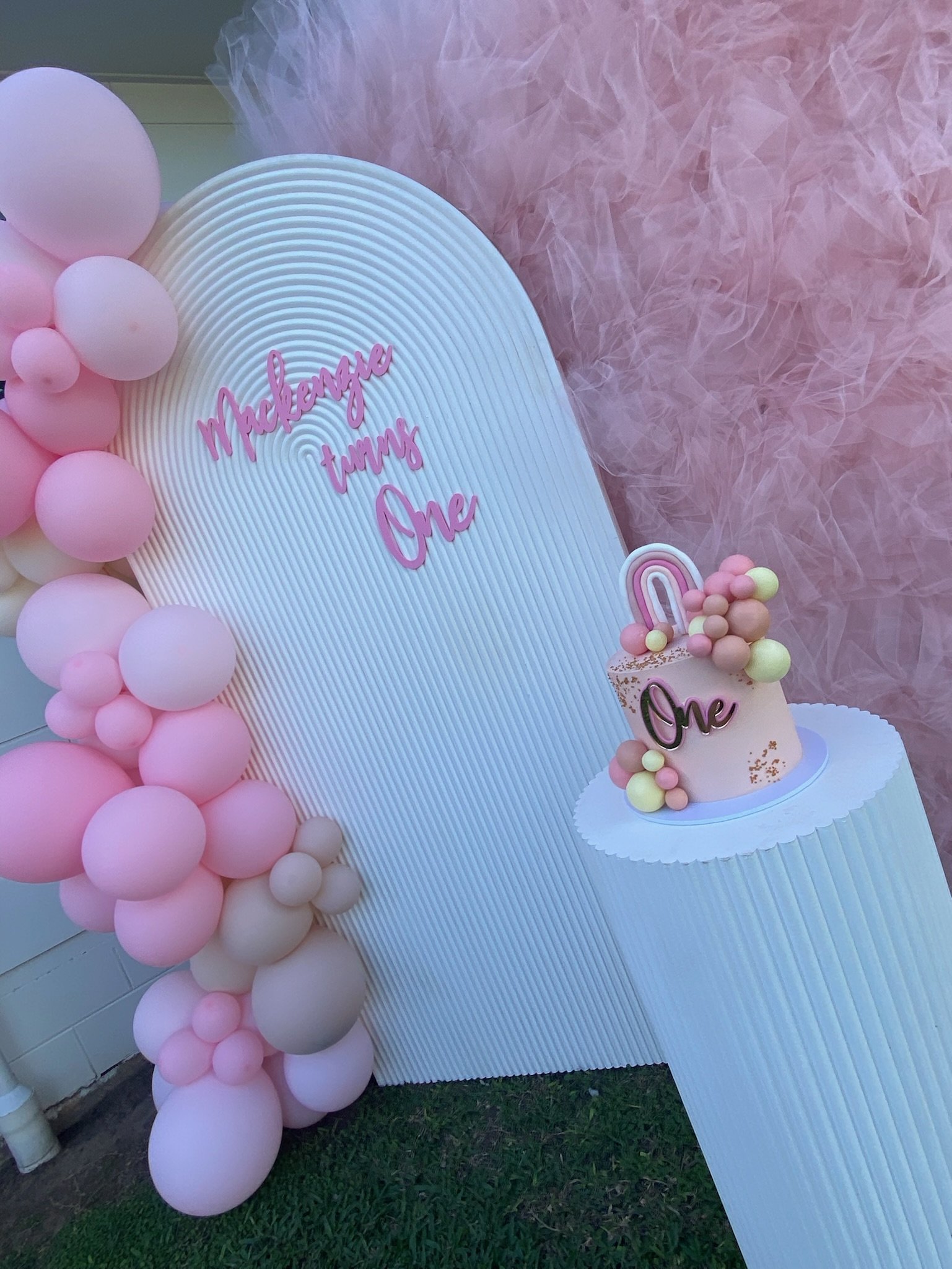 Decorative birthday display with pink and white balloons, a white textured backdrop, and a small pink cake with a rainbow and colored balls on top. The cake has a black 'One' topper, and a pink sign on the backdrop reads 'Mackenzie turns One' in pink