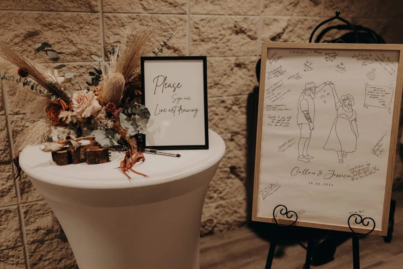 A wedding guestbook display with a framed illustration of a couple and handwritten messages for the bride and groom, with a floral arrangement on a white table.