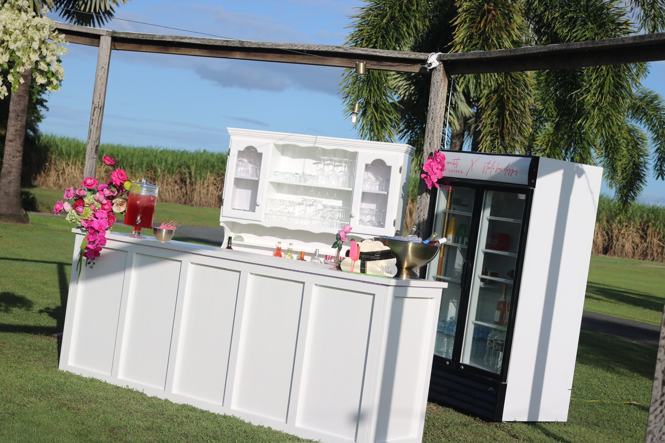 An outdoor bar setup with a white counter, glassware cabinet, and a refrigeration unit on grass with trees in the background under a blue sky.