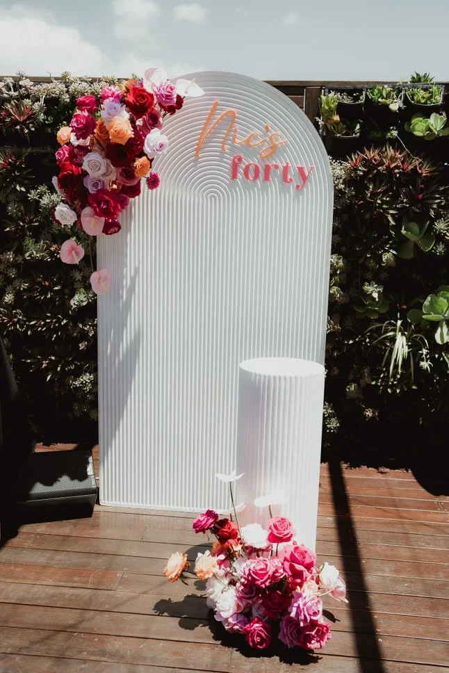 A white outdoor backdrop panel decorated with pink and red flowers, with the words 'Mrs forly' written in peach and pink. There are additional floral arrangements at the bottom and side of the panel, and a small white cylindrical stand in front, all 